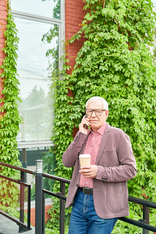 Portrait of modern senior businessman speaking by smartphone standing on terrace outdoors, copy space