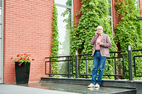 Full length portrait of modern senior businessman speaking by smartphone standing on terrace outdoors and holding paper coffee cup, copy space