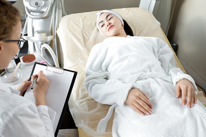 Unrecognizable young beautician wearing eyeglasses and white coat sitting in front of pretty client with clipboard in hands and giving her recommendations after completion of cosmetic procedure.