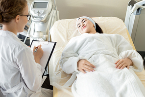 Portrait of young Asian woman lying on massage table and listening to cosmetologist giving instructions for treatment