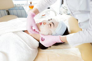 Head and shoulders portrait of pretty young woman lying on massage table enjoying beauty treatment with unrecognizable cosmetologist applying refreshing dace mask to her face