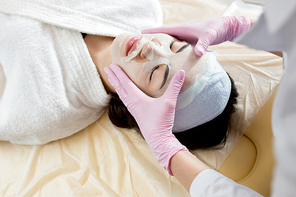 Unrecognizable beautician wearing rubber gloves applying sheet mask on face of her pretty young client, close-up shot