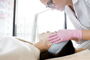 Side view portrait of pretty young woman lying on massage table enjoying beauty treatment with cosmetologist applying refreshing dace mask to her face