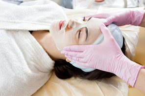 High angle portrait of pretty young woman lying on massage table enjoying beauty treatment with unrecognizable cosmetologist applying refreshing dace mask to her face