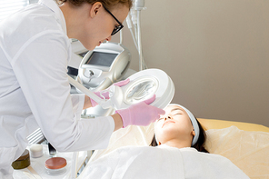 Portrait of  cosmetologist inspecting skin of young woman using magnifying lamp before beauty treatments