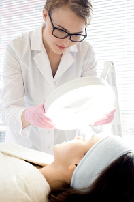 Concentrated young cosmetologist wearing eyeglasses and white coat looking through magnifying lamp at facial skin of young client while making preparations for procedure