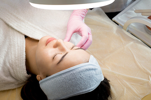 Portrait of pretty Asian woman with eyes closed laying under magnifying lamp in cosmetologist office, preparing for beauty treatment