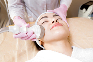 Headshot of pretty young woman wearing bathrobe and headband lying on treatment table of beauty salon while getting ultrasound face cleaning