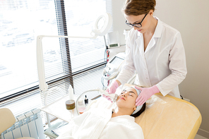 Relaxed young woman wearing bathrobe lying on treatment table while highly professional beautician massaging her face with special equipment, interior of modern facial room on background