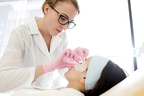 Side view portrait of female cosmetologist applying refreshing eye mask to face of beautiful young woman performing beauty treatment