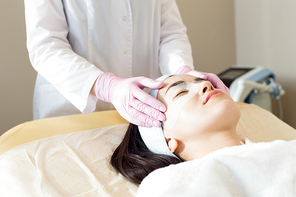 Close up of unrecognizable cosmetologist applying refreshing eye mask to face of beautiful young woman while performing beauty treatment