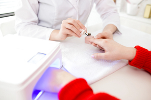 Close-up shot of unrecognizable manicurist wearing white coat covering nails of female client with gel polish while sitting opposite her