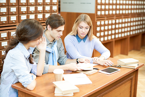 Group of serious concentrated student in casual clothing reading book together thoughtfully while preparing for exam in university library