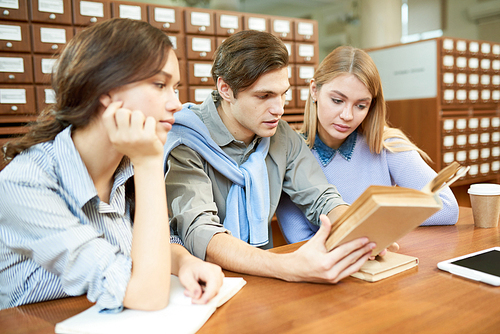 Two pretty students and their handsome groupmate gathered together in spacious library and studying hard, catalogue cabinets on background