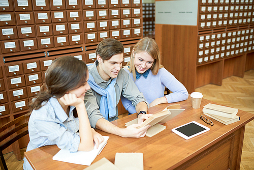 Positive excited young students sitting at table and studying textbook together to prepare for important exam in college library