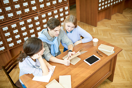 High angle view of hard-working group of university students sitting at wooden desk and doing their homework together, interior of spacious library on background