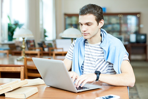 Serious concentrated handsome young student with sweater on shoulders typing on laptop while working on research and summarizing books in library