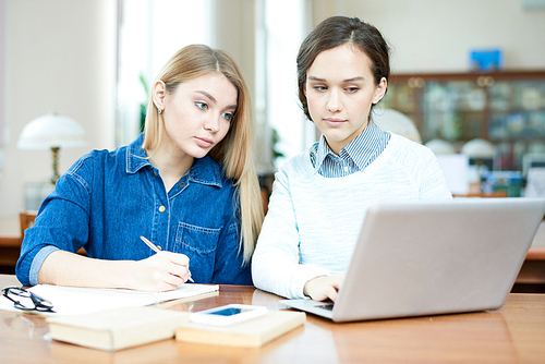 Attractive young friends gathered together in spacious library and doing university assignment together, dark-haired girl using laptop