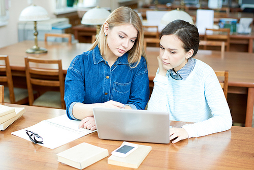 Pretty blond-haired college student wearing denim shirt sitting at wooden table with her groupmate and sharing ideas concerning their joint project