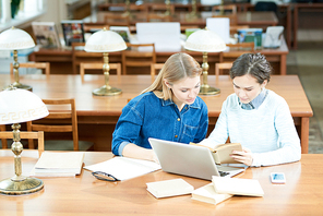 Busy concentrated modern student girl analyzing textbook and using laptop while sitting at table in cozy old-fashioned library