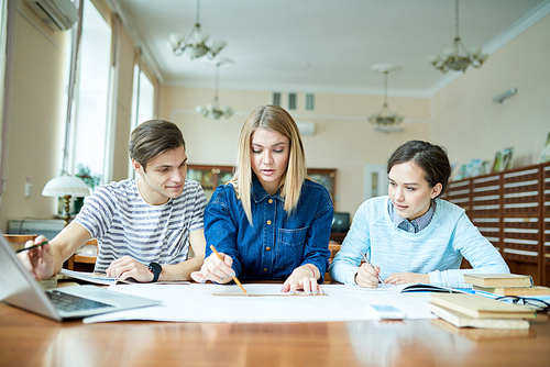 Group of hard-working college students studying hard for exam while sitting at desk of spacious library,  catalogue cabinets seen behind them