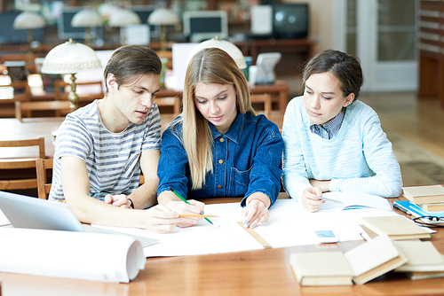 Group of pensive concentrated architecture students discussing blueprint while sitting at table in library