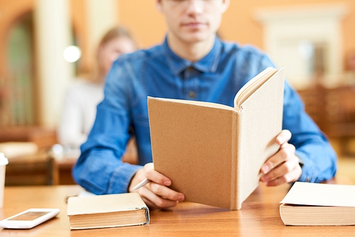 Close-up shot of unrecognizable library visitor sitting at wooden table and reading book, blurred background