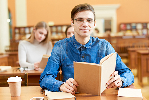 Portrait of pleased confident handsome young male student holding open textbook and sitting at table in library
