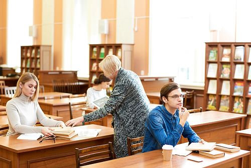 Attractive young woman sitting at wooden desk of spacious library while blond-haired librarian consulting her about availability of necessary book, other students focused on their assignments