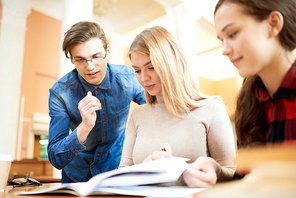 Positive confident handsome young male student explaining difficult topic to concentrated girls in modern library while they studying material together