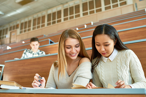 Portrait of two beautiful girls enjoying studying sitting at desk in lecture hall of modern college and taking notes, copy space