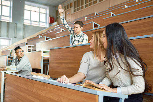 Multi-ethnic group of people sitting at separate tables in lecture hall of modern college, focus on two beautiful girls in foreground turning to look at young man raising hand in class