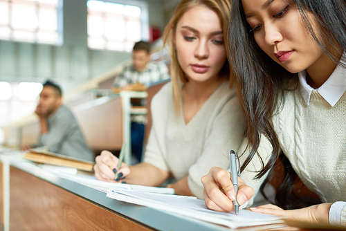 Portrait of two beautiful young women sitting at desk in lecture hall of modern university and writing notes, copy space