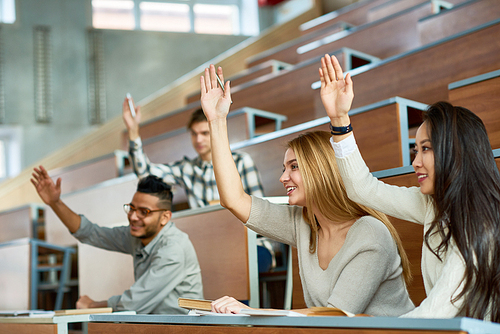 Multi-ethnic group of students sitting at separate tables in lecture hall of modern college and raising hands, focus on two beautiful girls  smiling