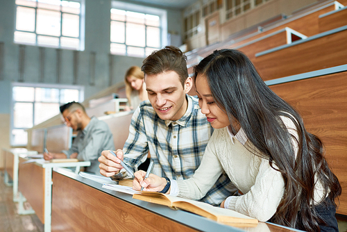 Portrait of two cheerful students sitting at desk in lecture hall of modern college, young man and Asian woman enjoying class, copy space