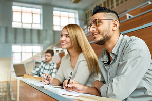 Multi-ethnic group of students sitting at desk in lecture hall of modern college and smiling happily, focus on young Middle-Eastern man wearing glasses, copy space