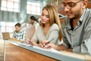 Multi-ethnic group of students sitting at desk in lecture hall of modern college and writing, focus on young Middle-Eastern man wearing glasses, copy space