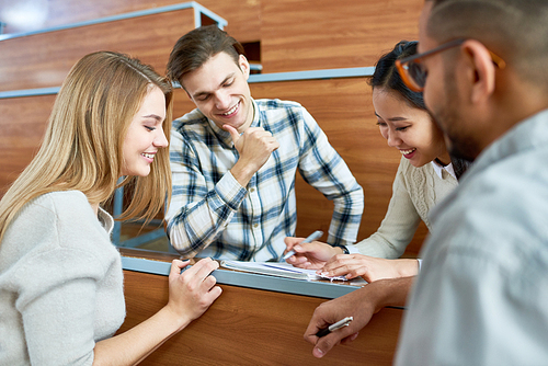 Group of international students sitting around desk in modern lecture hall and studying together, smiling happily and making notes