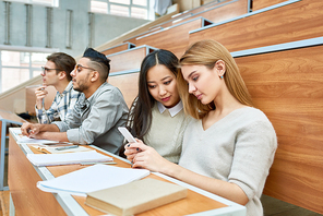 International group of students sitting in row at desks in modern lecture hall waiting for class in modern college, focus on two pretty girls using smartphone