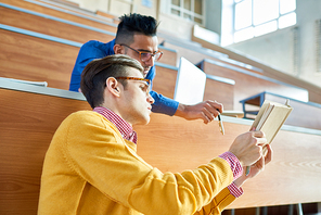 Portrait of two students discussing lecture sitting in auditorium before class, focus on young man wearing glasses showing open book to Middle-Eastern student