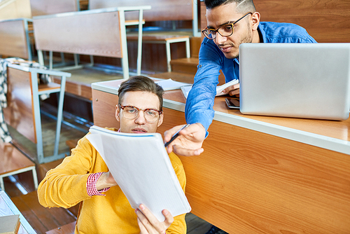 Portrait of two students discussing lecture sitting in auditorium before class, focus on young man wearing glasses showing notes to Middle-Eastern student