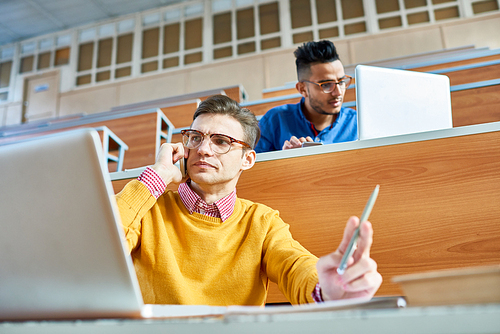 Low angle view of two students, one of them Middle-eastern, sitting at desks in modern auditorium at college and preparing for class, focus on young man using laptop and speaking by smartphone