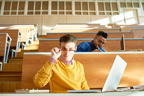 Portrait of two students, one of them Middle-eastern, sitting at desks in modern auditorium at college and preparing for class, focus on smart young man using laptop and 