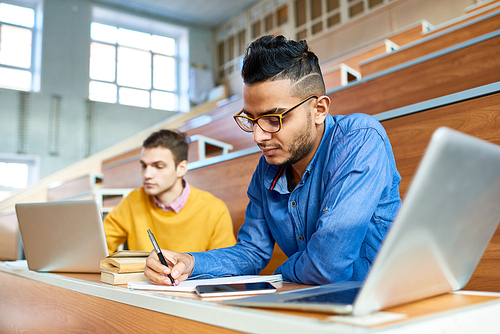 Portrait of two students sitting at desks in modern auditorium at college and preparing for class, focus on young Middle-Eastern man writing in copybook