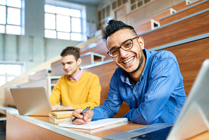 Portrait of two students sitting at desks in modern auditorium at college and preparing for class, focus on young Middle-Eastern man  and smiling, copy space