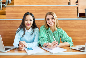 Portrait of two pretty girls in college, one of them Asian, smiling ad  while sitting at desk in modern lecture hall