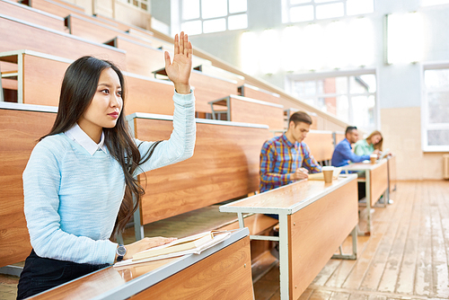 Portrait of beautiful Asian woman raising hand during class at college, copy space with people in background