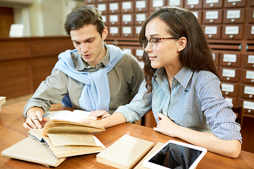 Group of hard-working students writing report together while sitting at wooden desk of reading room, catalogue cabinets on background