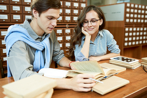 Smiling content confident students preparing for important exam surrounded by textbooks, they sitting at table and reading book in library