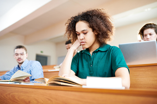 Serious concentrated attractive African-American student girl with Afro hairstyle leaning on hand and reading textbook while sitting at university desk in lecture room together groupmates.